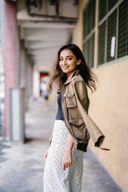 Portrait of a tall, beautiful, slim and elegant Indian Asian woman walking down a walkway in a city in Asia during the day. She is elegantly dressed and has a jacket draped over her shoulders. clipart