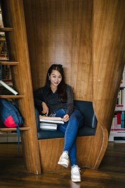 Genç, güzel ve çekici Çinli Asyalı değişim öğrencisi kütüphanedeki kahverengi bir kulübede okuyor ve çalışıyor..