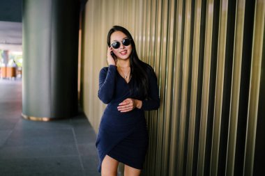 Modern bir altın özelliği duvara karşı şık ve zarif mavi elbise genç ve güzel Asyalı Çinli kadın Portresi. Akıllı telefonuyla konuşurken havalı güneş gözlüğü takıyor.