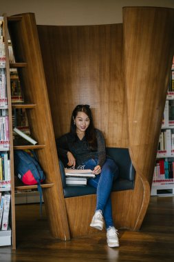 Genç, güzel ve çekici Çinli Asyalı değişim öğrencisi kütüphanedeki kahverengi bir kulübede okuyor ve çalışıyor..