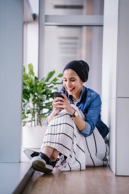 Genç bir Ortadoğulu Müslüman kadın gün boyunca bir ofiste, ortak çalışma alanında veya kütüphanede oturuyor ve akıllı telefonunu kullanıyor. Başörtüsü takıyor (başörtüsü)).
