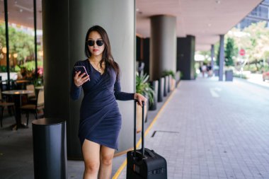 O bir yolculuk dolu uygulama üzerinden rezervasyonu onu binmek için caddenin kenarında bekleyen zarif ve güzel bir genç Asyalı kadının portresi. Tekerlekli bir bavula tutunuyor.. 