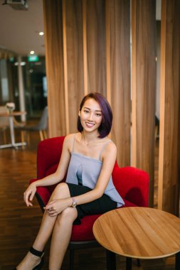 Profesyonel elbiseli ve boyalı mor saçlı genç, ince ve zarif bir Asyalı kadın çalışma odasında kırmızı bir koltukta oturuyor. İçtenlikle gülümsüyor..