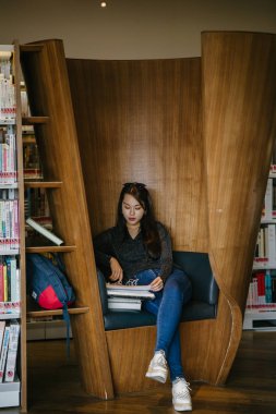 Genç, güzel ve çekici Çinli Asyalı değişim öğrencisi kütüphanedeki kahverengi bir kulübede okuyor ve çalışıyor..