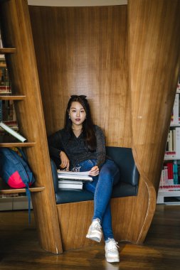 Genç, güzel ve çekici Çinli Asyalı değişim öğrencisi kütüphanedeki kahverengi bir kulübede okuyor ve çalışıyor..