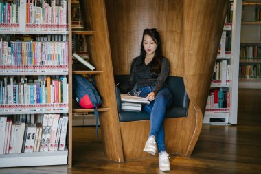 Genç, güzel ve çekici Çinli Asyalı değişim öğrencisi kütüphanedeki kahverengi bir kulübede okuyor ve çalışıyor..