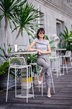 Açık havada şık bir yerde oturan bir emin, şık ve çekici orta yaşlı Çinli Asyalı kadın portresi. O bir pohpohlama gri kıyafeti giyiyor.