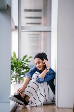 Genç bir Ortadoğulu Müslüman kadın gün boyunca bir ofiste, ortak çalışma alanında veya kütüphanede oturuyor ve akıllı telefonunu kullanıyor. Başörtüsü takıyor (başörtüsü)).