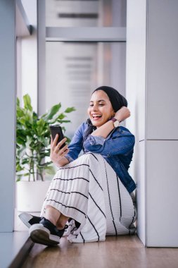 Genç bir Ortadoğulu Müslüman kadın gün boyunca bir ofiste, ortak çalışma alanında veya kütüphanede oturuyor ve akıllı telefonunu kullanıyor. Başörtüsü takıyor (başörtüsü)).