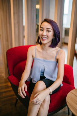 Profesyonel elbiseli ve boyalı mor saçlı genç, ince ve zarif bir Asyalı kadın çalışma odasında kırmızı bir koltukta oturuyor. İçtenlikle gülümsüyor..