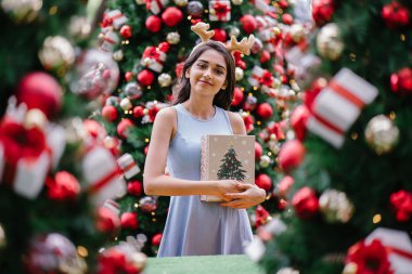 Genç, güzel ve mutlu bir Hint Asyalı kadın şenlikli Noel portresi bir hediye bir Noel ağacı önünde tutan gülümseyerek.