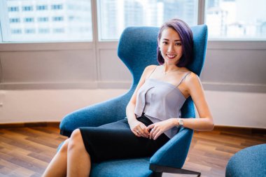 Profesyonel elbiseli ve boyalı mor saçlı genç, ince ve zarif bir Asyalı kadın çalışma odasında kırmızı bir koltukta oturuyor. İçtenlikle gülümsüyor..