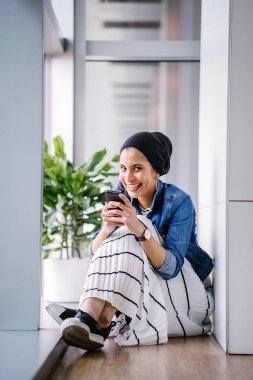 Genç bir Ortadoğulu Müslüman kadın gün boyunca bir ofiste, ortak çalışma alanında veya kütüphanede oturuyor ve akıllı telefonunu kullanıyor. Başörtüsü takıyor (başörtüsü)).