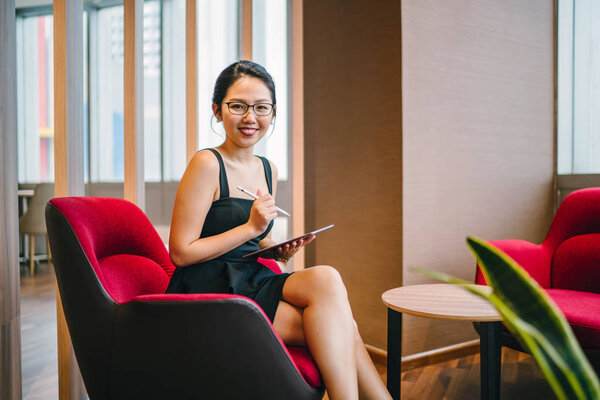 A business portrait of a young and beautiful Asian woman working on her tablet device with a stylus pen as she sits in her coworking office during the day.