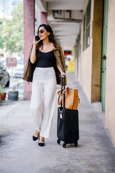 young and attractive Asian Indian woman books a ride through her ride hailing app on her smartphone. She is standing in a walkway with her luggage and is stylishly dressed in work wear and shades