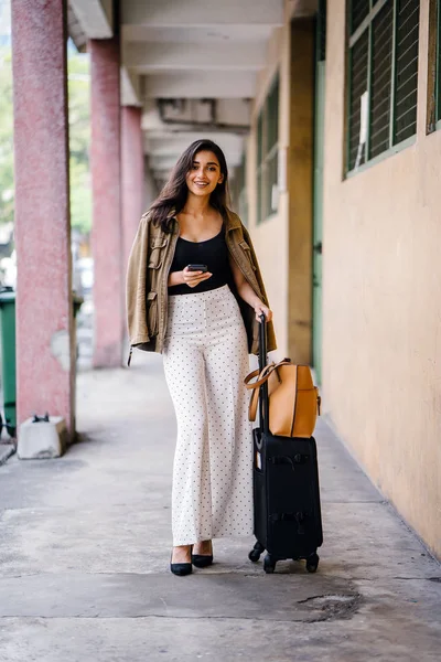 young and attractive Asian Indian woman books a ride through her ride hailing app on her smartphone. She is standing in a walkway with her luggage and is stylishly dressed in work wear and shades