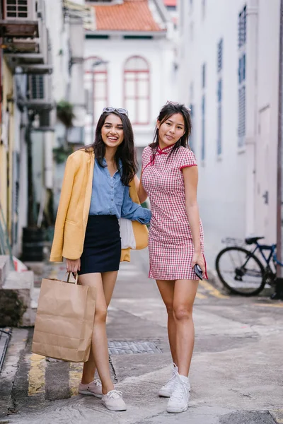 two close friends walking together down an alley. They are both wearing trendy retro clothing and holding hands as they walk happily together. One is Chinese, the other Indian