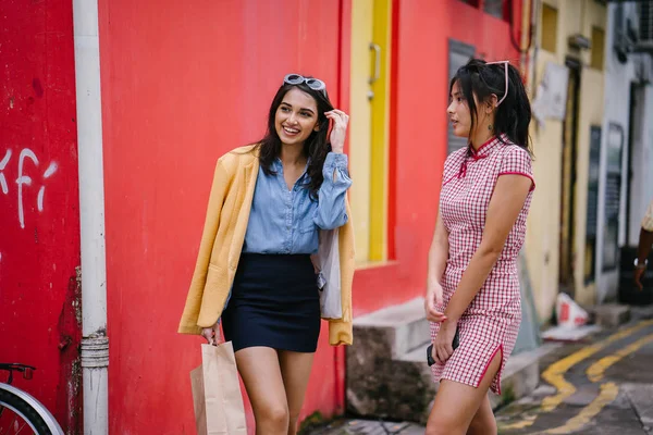 two close friends walking together down an alley. They are both wearing trendy retro clothing and holding hands as they walk happily together. One is Chinese, the other Indian