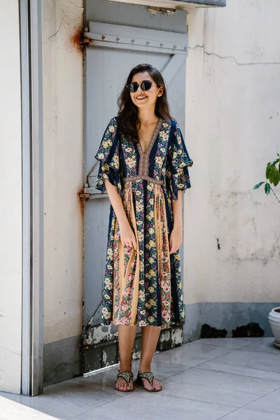 tall, elegant, and beautiful Indian Asian woman in a summer dress and sunglasses smiles as she poses on a street in an alley during the day