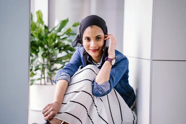 A young Middle-Eastern Muslim woman sits in a corner of an office, coworking space or library during the day and is using her smartphone. She is wearing a head scarf (hijab).