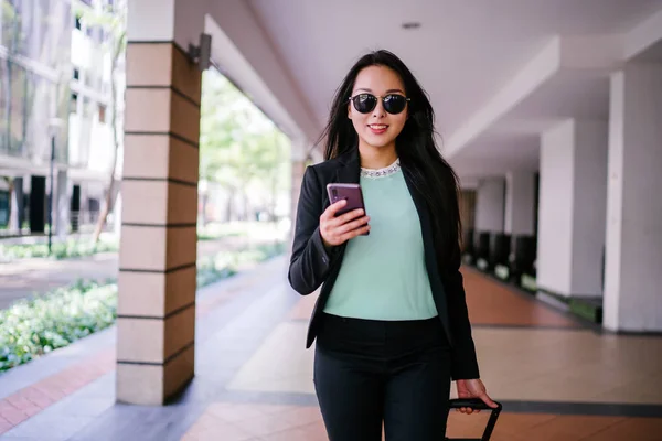 Portrait of an elegant and beautiful young Asian woman waiting on the side of the street for her ride that she booked via a ride-hailing app. She is holding onto a luggage suitcase on wheels.