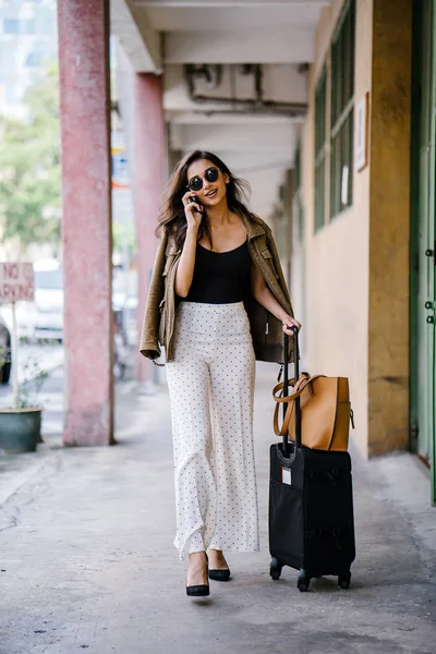 young and attractive Asian Indian woman books a ride through her ride hailing app on her smartphone. She is standing in a walkway with her luggage and is stylishly dressed in work wear and shades