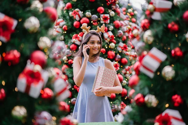 Genç, güzel ve mutlu bir Hint Asyalı kadın şenlikli Noel portresi bir hediye bir Noel ağacı önünde tutan gülümseyerek.