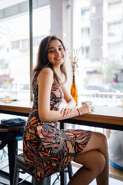 Young and beautiful Indian Asian Singaporean woman in a print dress sits in a cafe as she enjoys the weekend in Asia.