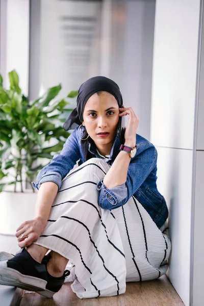 一名年轻的中东穆斯林妇女白天坐在办公室 工作空间或图书馆的角落里 用她的智能手机 她戴着头巾 — 图库照片
