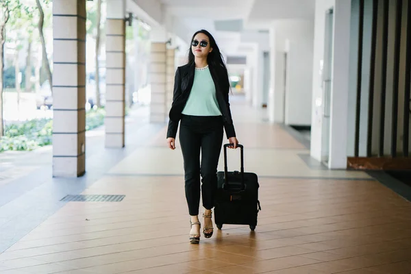 Portrait of an elegant and beautiful young Asian woman waiting on the side of the street for her ride that she booked via a ride-hailing app. She is holding onto a luggage suitcase on wheels.