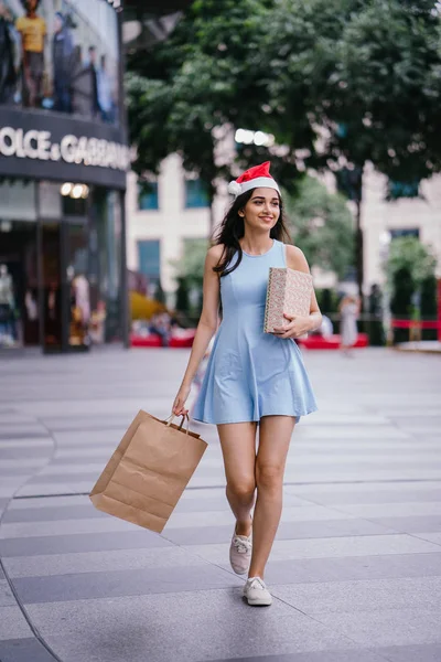商店街での買い物袋と歩いている若い かわいいインド アジア少女のクリスマスの肖像画 彼女は赤いクリスマス サンタ帽子をかぶっては 彼女は彼女のバッグを持って歩く笑みを浮かべて — ストック写真