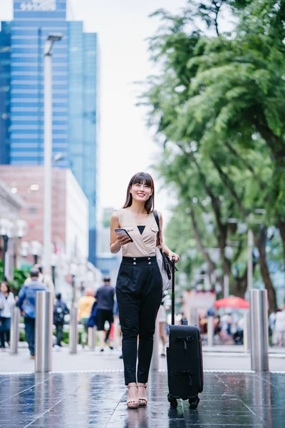 attractive Asian Chinese woman books a ride through her ride hailing app on her smartphone. She is walking with her luggage and is stylishly dressed in work wear