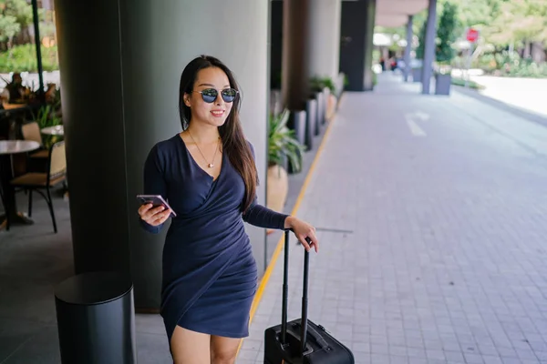 Portrait of an elegant and beautiful young Asian woman waiting on the side of the street for her ride that she booked via a ride-hailing app. She is holding onto a luggage suitcase on wheels.