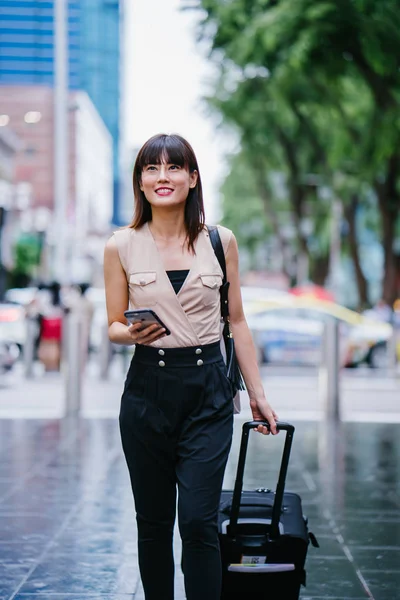 attractive Asian Chinese woman books a ride through her ride hailing app on her smartphone. She is walking with her luggage and is stylishly dressed in work wear