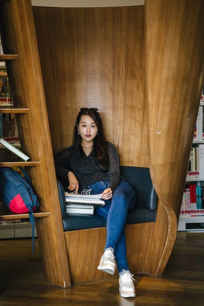 Genç, güzel ve çekici Çinli Asyalı değişim öğrencisi kütüphanedeki kahverengi bir kulübede okuyor ve çalışıyor..