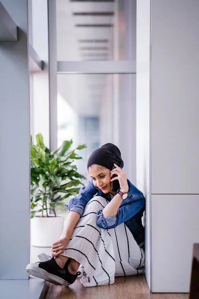 A young Middle-Eastern Muslim woman sits in a corner of an office, coworking space or library during the day and is using her smartphone. She is wearing a head scarf (hijab).