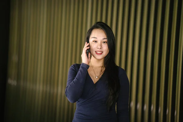 一个年轻漂亮的亚洲中国妇女的肖像 穿着优雅优雅的蓝色连衣裙 与现代金色特色墙 她穿着很酷的太阳镜 在她的智能手机上说话 — 图库照片