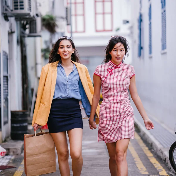 two close friends walking together down an alley. They are both wearing trendy retro clothing and holding hands as they walk happily together. One is Chinese, the other Indian