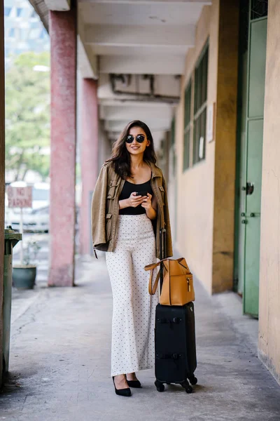 young and attractive Asian Indian woman books a ride through her ride hailing app on her smartphone. She is standing in a walkway with her luggage and is stylishly dressed in work wear and shades