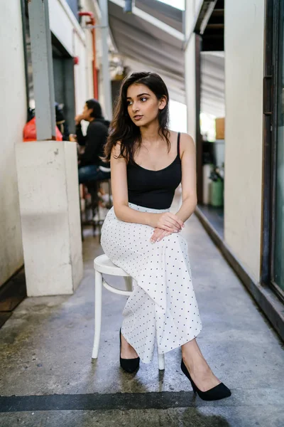 Bir uzun boylu, zarif ve güzel Hint gün boyunca şehrin bir kafede bir sandalyede oturan kız en iyi ve uzun beyaz siyah pantolon Asya portresi
