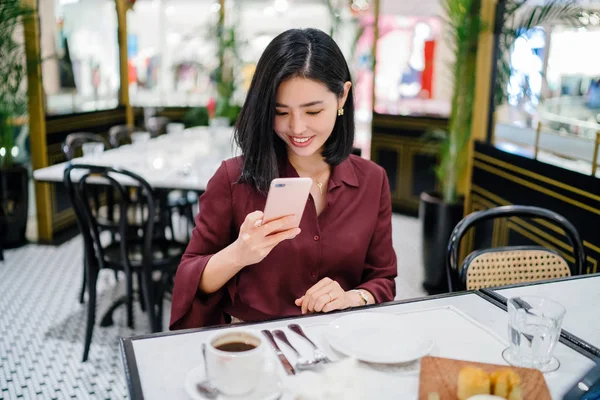 Şık bir kapalı kafede oturan ve akıllı telefon tutan güzel ve zarif genç Koreli Asyalı kadının bir portre