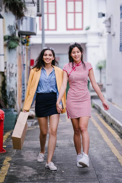 two close friends walking together down an alley. They are both wearing trendy retro clothing and holding hands as they walk happily together. One is Chinese, the other Indian