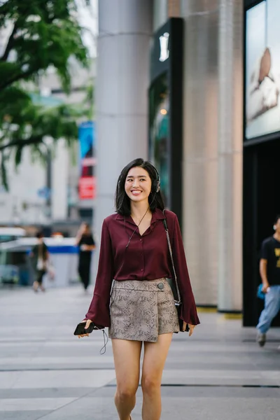 beautiful Korean woman in an elegant blouse and shorts dancing as she walks down a street in the city with her headphones on