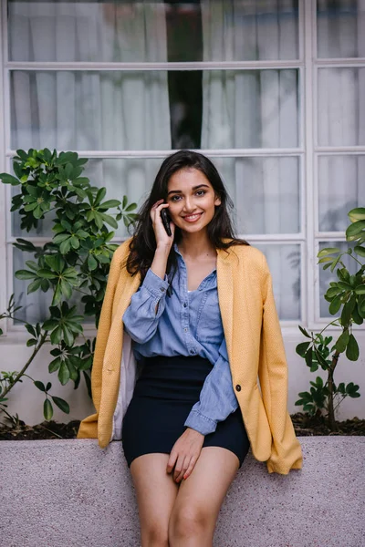 güzel, genç ve şık Hint Asyalı kadın o gün bir çıkıntı karşı eğilir gibi onun smartphone üzerinde söz. O rahat kıyafetler üzerinde bir retro sarı ceket giyiyor