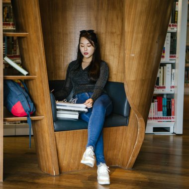 Genç, güzel ve çekici Çinli Asyalı değişim öğrencisi kütüphanedeki kahverengi bir kulübede okuyor ve çalışıyor..