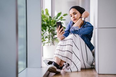 Genç bir Ortadoğulu Müslüman kadın gün boyunca bir ofiste, ortak çalışma alanında veya kütüphanede oturuyor ve akıllı telefonunu kullanıyor. Başörtüsü takıyor (başörtüsü)).