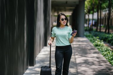 O bir yolculuk dolu uygulama üzerinden rezervasyonu onu binmek için caddenin kenarında bekleyen zarif ve güzel bir genç Asyalı kadının portresi. Tekerlekli bir bavula tutunuyor.. 