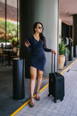 O bir yolculuk dolu uygulama üzerinden rezervasyonu onu binmek için caddenin kenarında bekleyen zarif ve güzel bir genç Asyalı kadının portresi. Tekerlekli bir bavula tutunuyor.. 