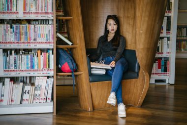 Genç, güzel ve çekici Çinli Asyalı değişim öğrencisi kütüphanedeki kahverengi bir kulübede okuyor ve çalışıyor..