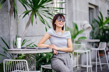 Açık havada şık bir yerde oturan bir emin, şık ve çekici orta yaşlı Çinli Asyalı kadın portresi. O bir pohpohlama gri kıyafeti giyiyor.